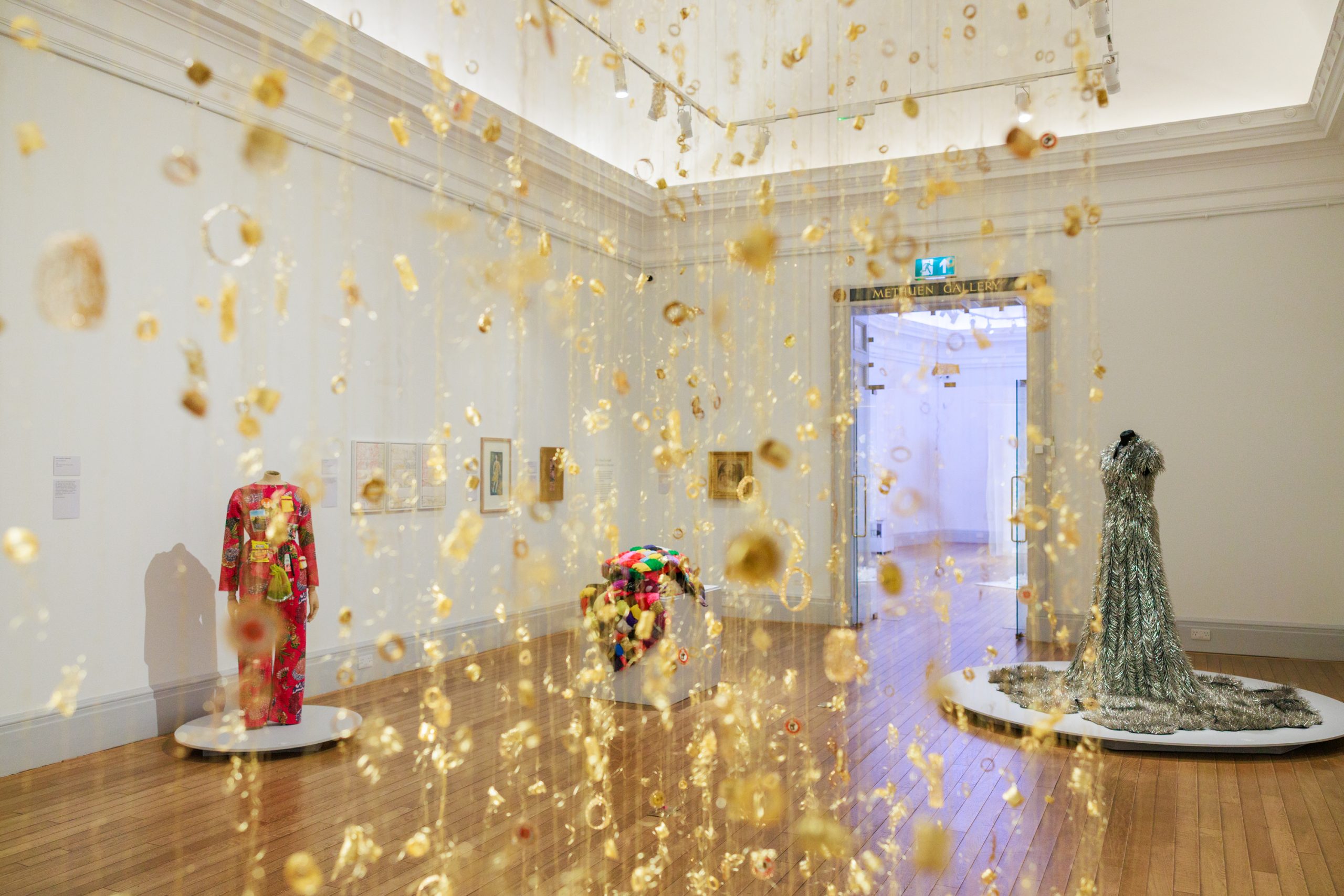 A close up shot of a hanging gold, delicate sculpture, between the dangling rows we see a larger exhibition space behind.