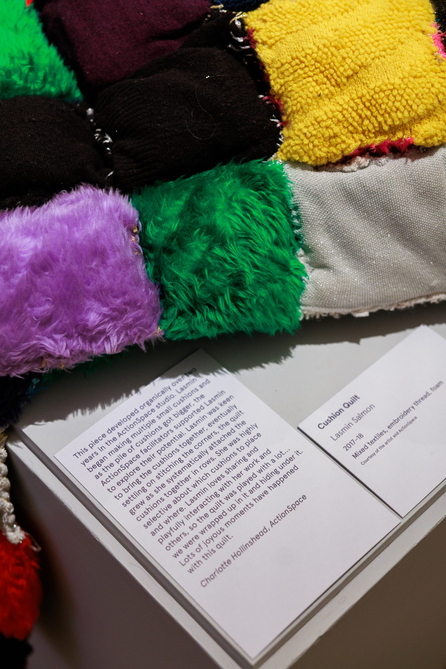 A close up shot of colourful, furry, textile patches, laid across a white surface with exhibition text in black on white panel.