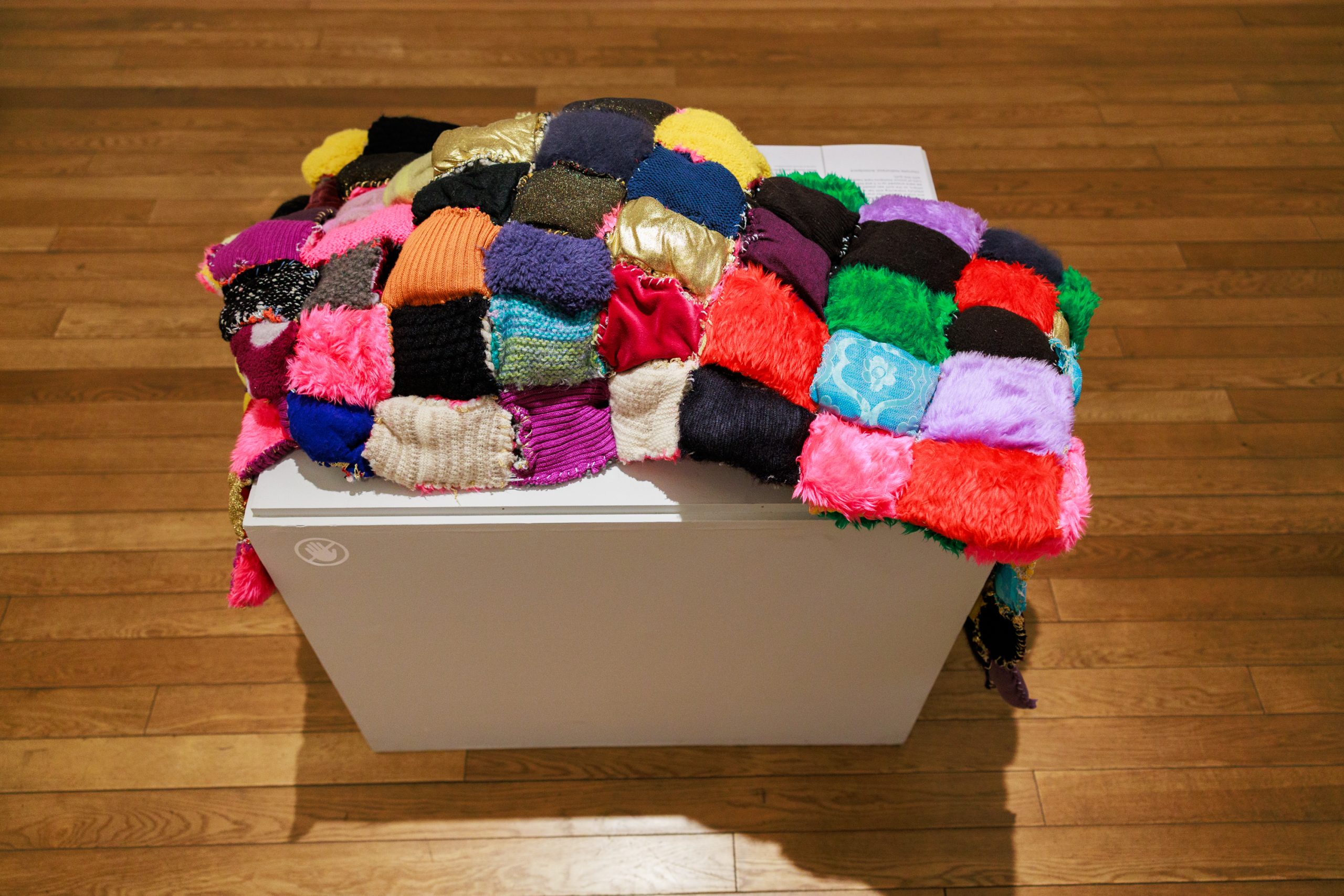 A colourful, patchwork fabric artwork laid on a white plinth on a brown, wooden floor.