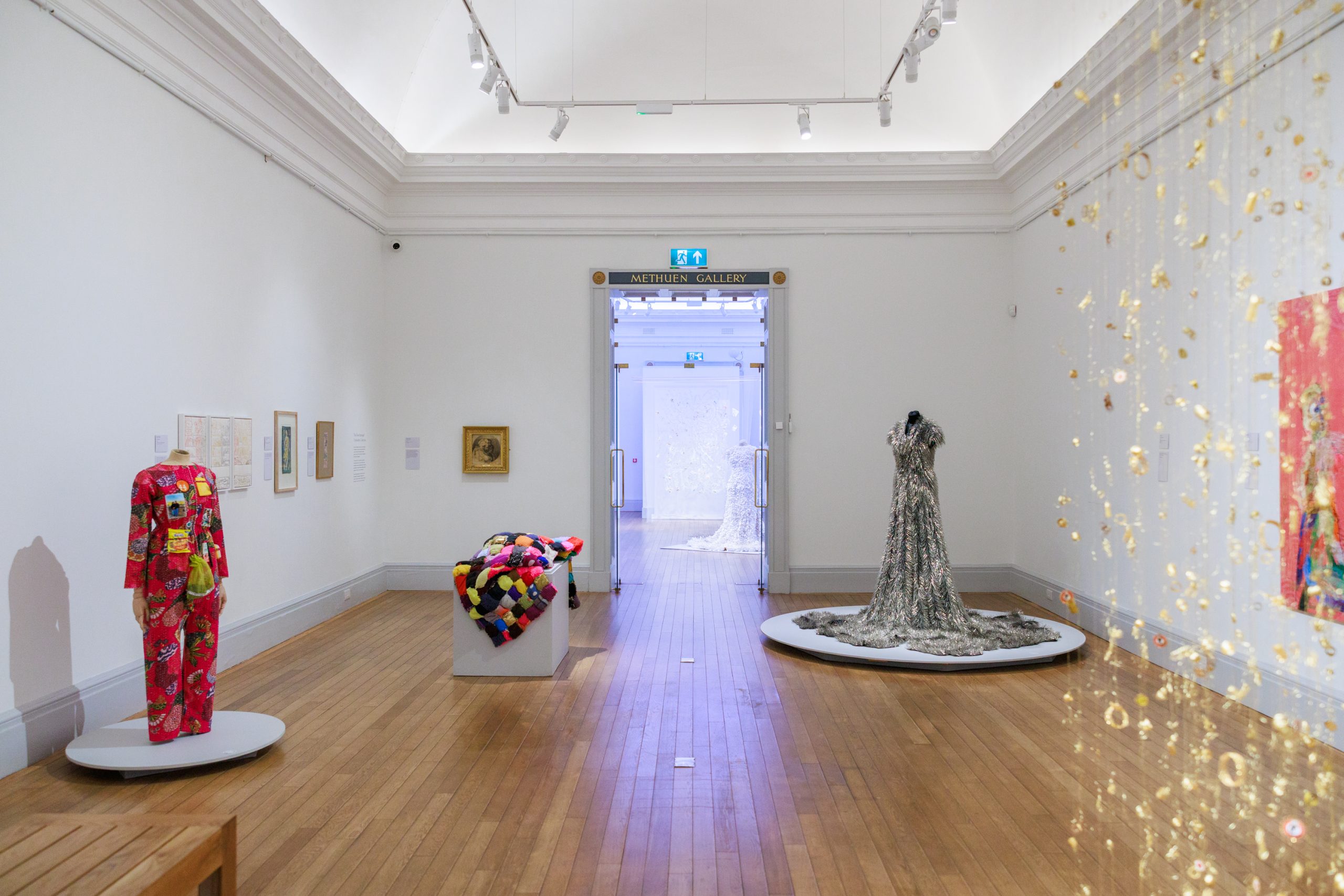 A large white gallery space with white walls and brown wooden floors, presenting a group exhibit of textile works carefully placed in the room.