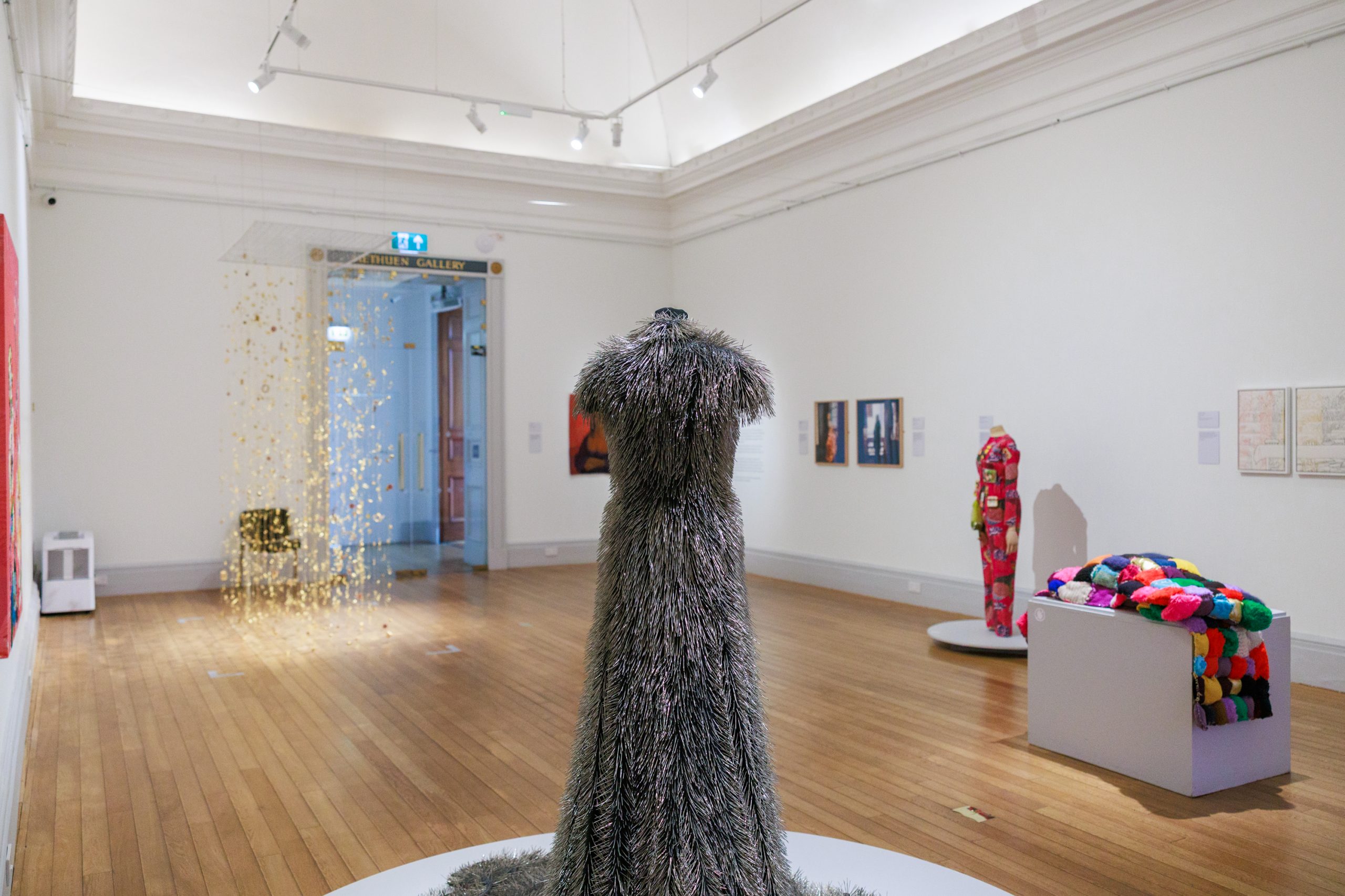 A large white gallery space with white walls and brown wooden floors, presenting a group exhibit of textile works carefully placed in the room.