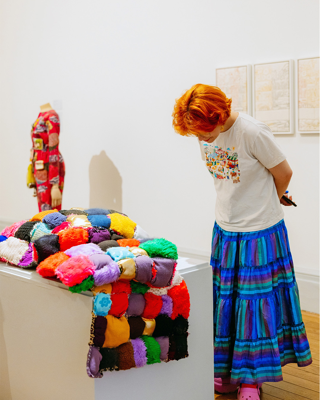 A white woman with a ginger bob looks down to observe a colourful, patchwork fabric artwork laid on a white plinth in a gallery.