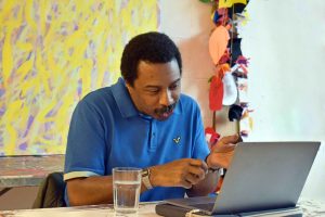 Black male artist sits at a table talking to the camera enthusiastically on his laptop.
