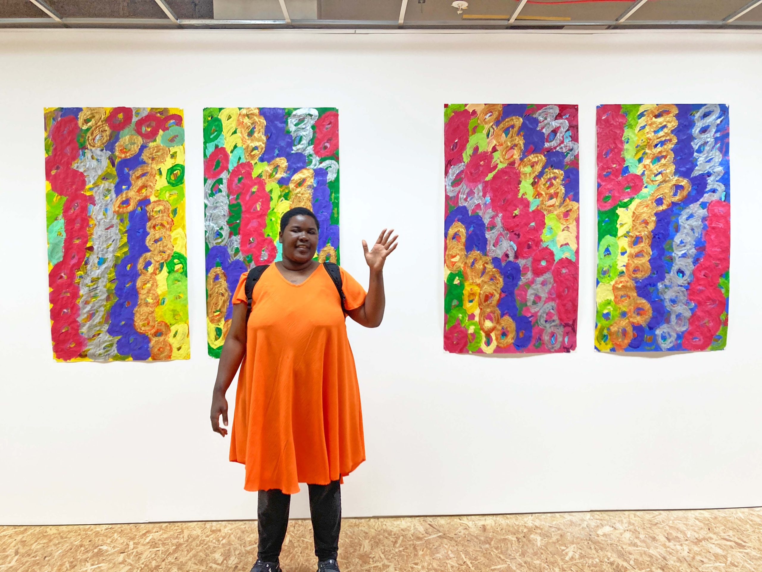 A female artist wearing an orange dress and black leggings waves to the camera in front of her artworks installed in a gallery.