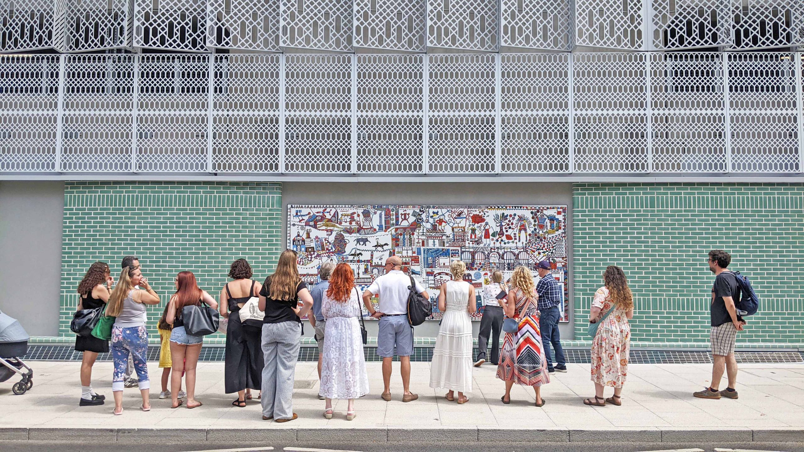A landscape image of a group of people looking at a large scale drawing installed on an exterior wall on a public street. The work is colourful and graphic. The sun shines.