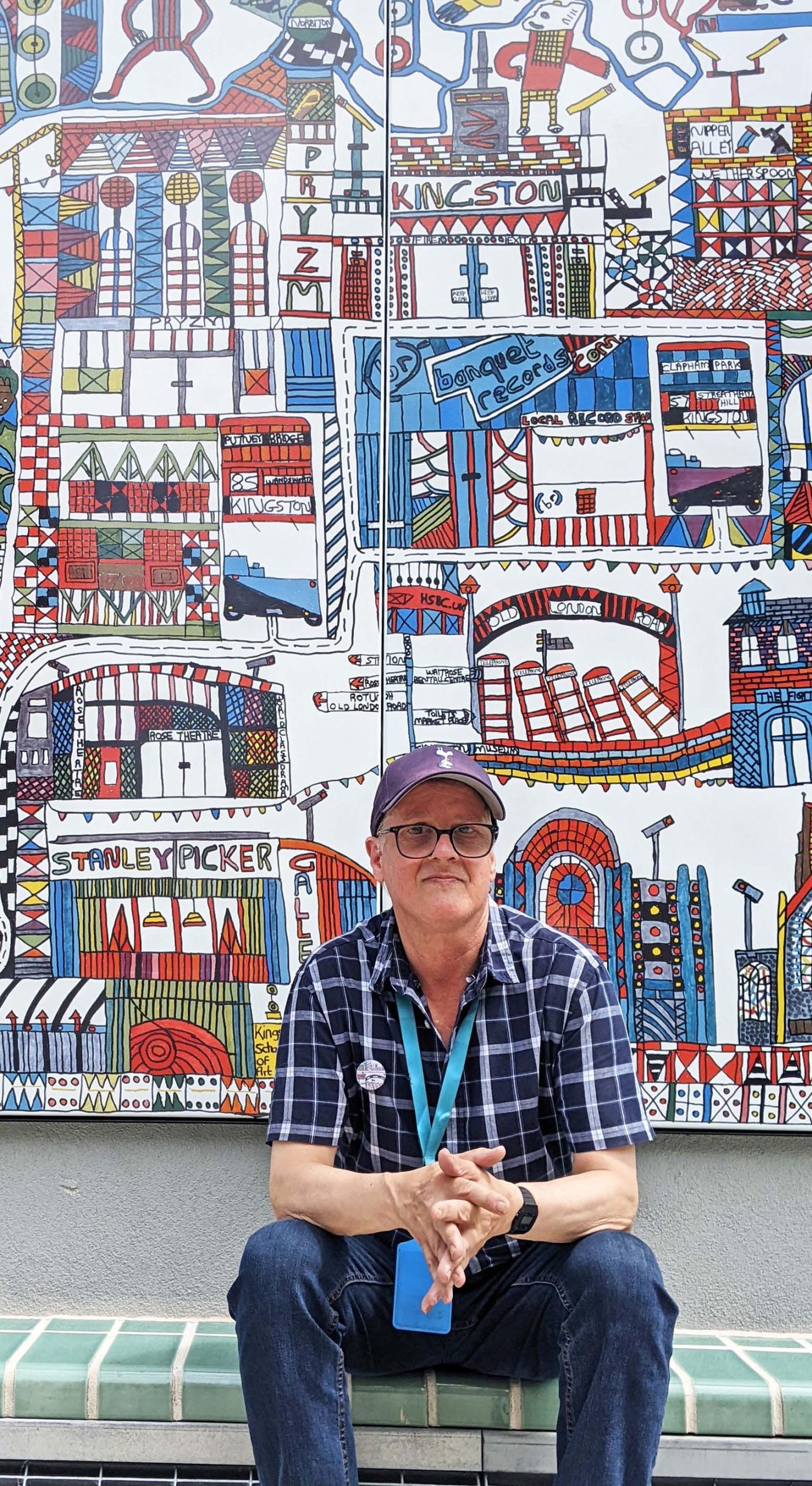 A white male artist wearing a cap and black glasses sits in front of his large scale installation outside. The work is graphic with colourful symbolism.