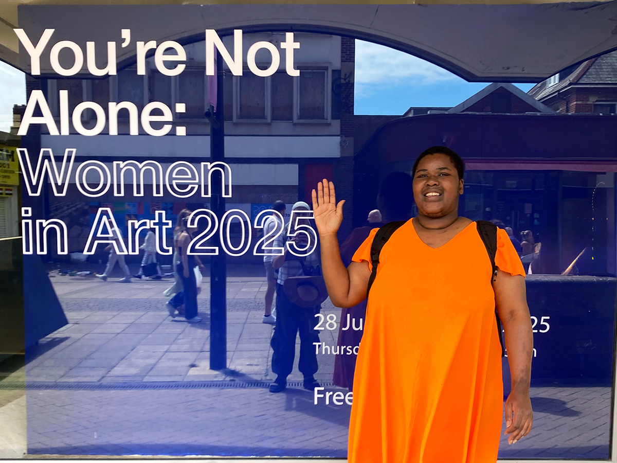 A black female artist in an orange long top waves to the camera in front of a blue gallery entrance.