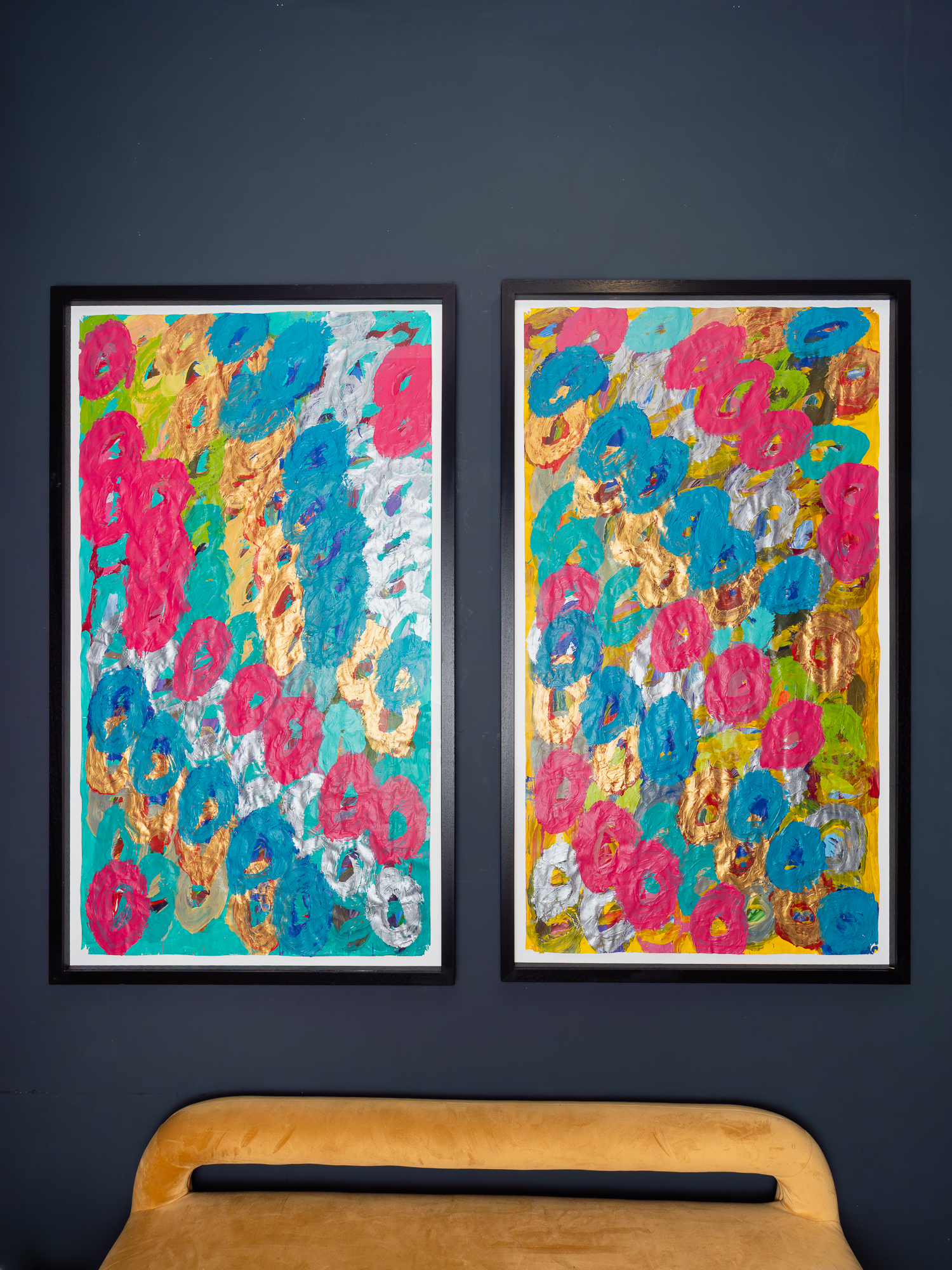 Image of two framed artworks hung straight, vertically on a blank black wall. The two colourful acrylic painting on large, white paper. A velvet mustard sofa sits beneath.