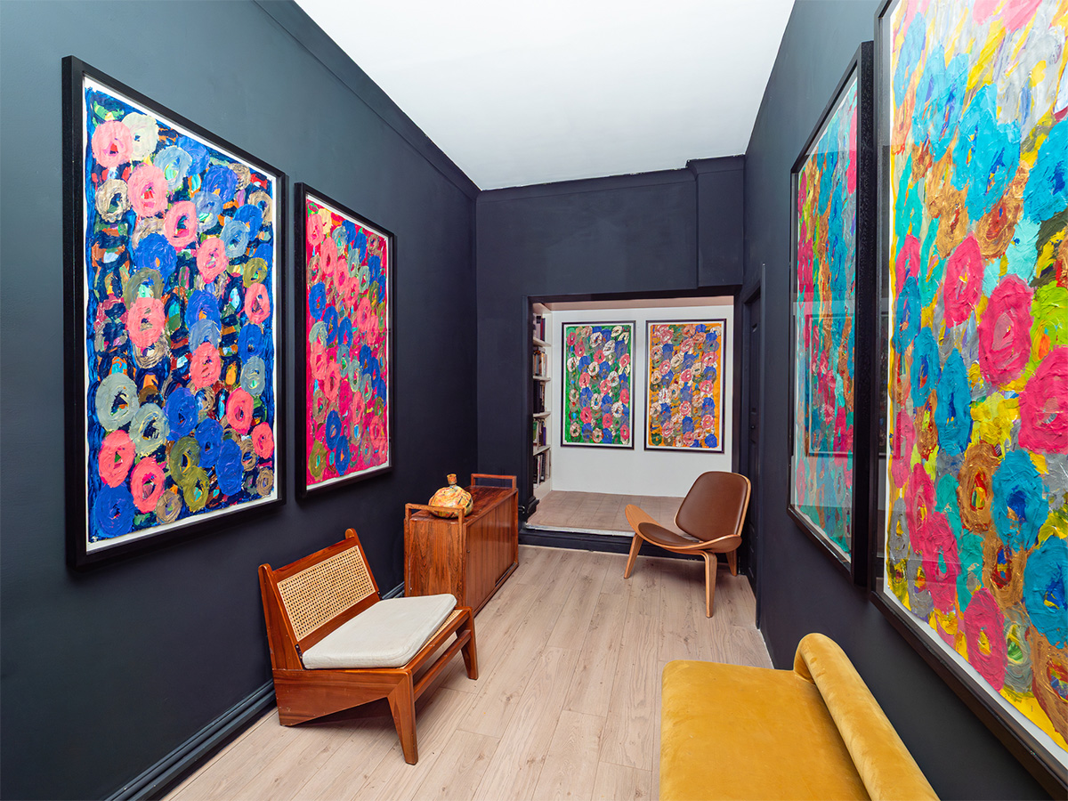 An image of a room with a white ceiling, brown wooden flooring, black walls, mid-century furnishing, and 6 large framed paintings hung in pairs on three walls. 