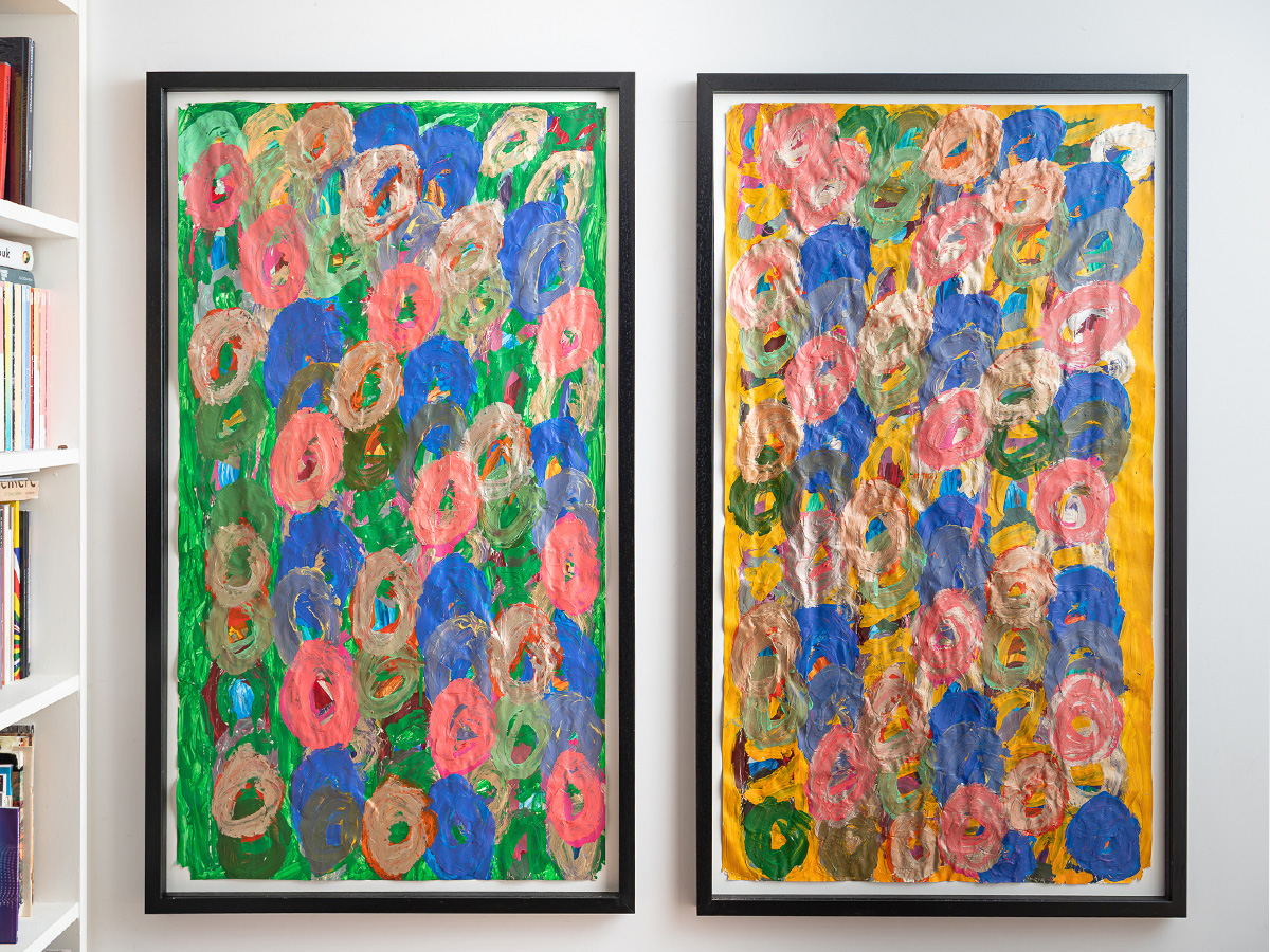 Image of two framed artworks hung straight, vertically on a blank white wall. The black frame displays a colourful acrylic painting on large paper with layers of sweeping paint. 