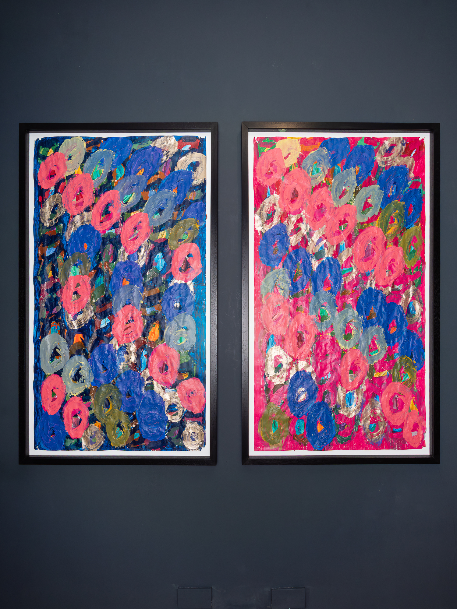 Image of two framed artworks hung straight, vertically on a blank black wall. The two colourful acrylic painting on large, white paper with layers of sweeping paint. 