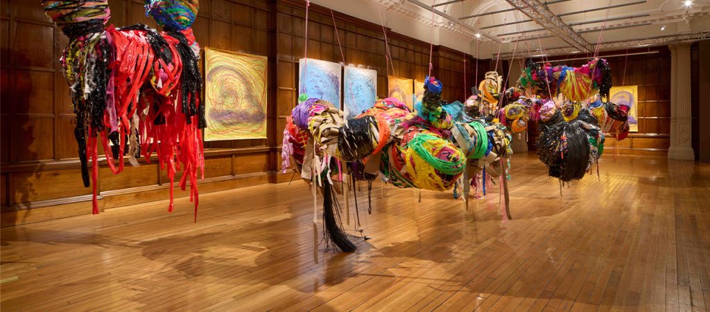 Installation view of various large mixed media sculptures suspended from the ceiling and swooping pencil drawings hung on the walls behind.