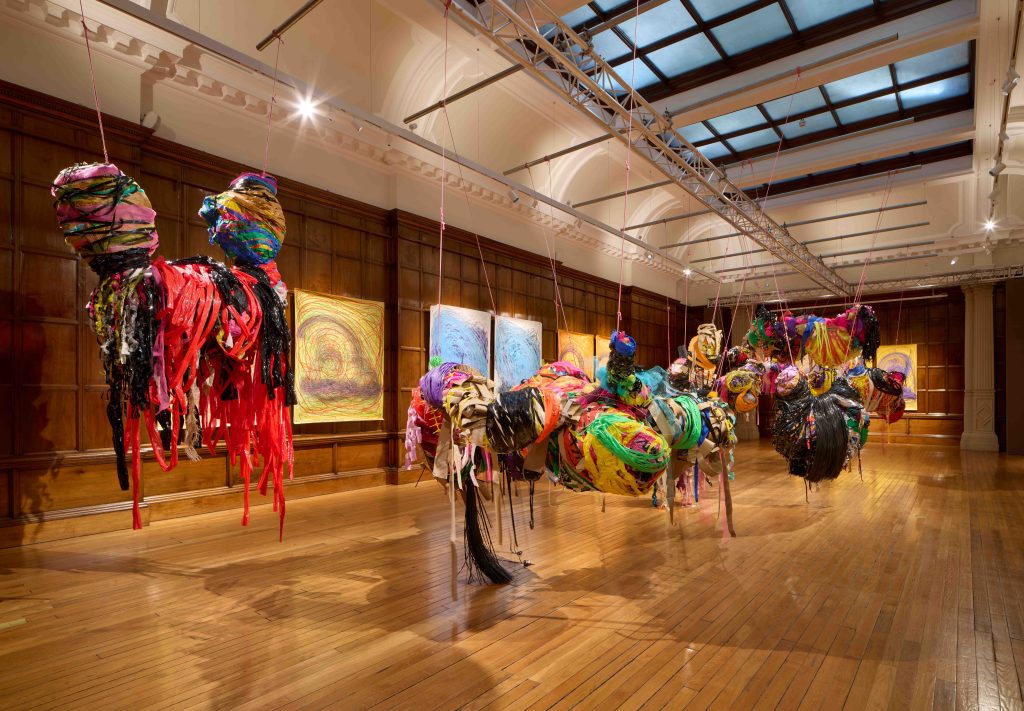 Installation view of various large mixed media sculptures suspended from the ceiling and swooping pencil drawings hung on the walls behind.