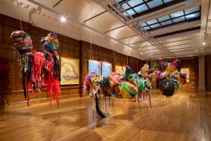Installation view of various large mixed media sculptures suspended from the ceiling and swooping pencil drawings hung on the walls behind.
