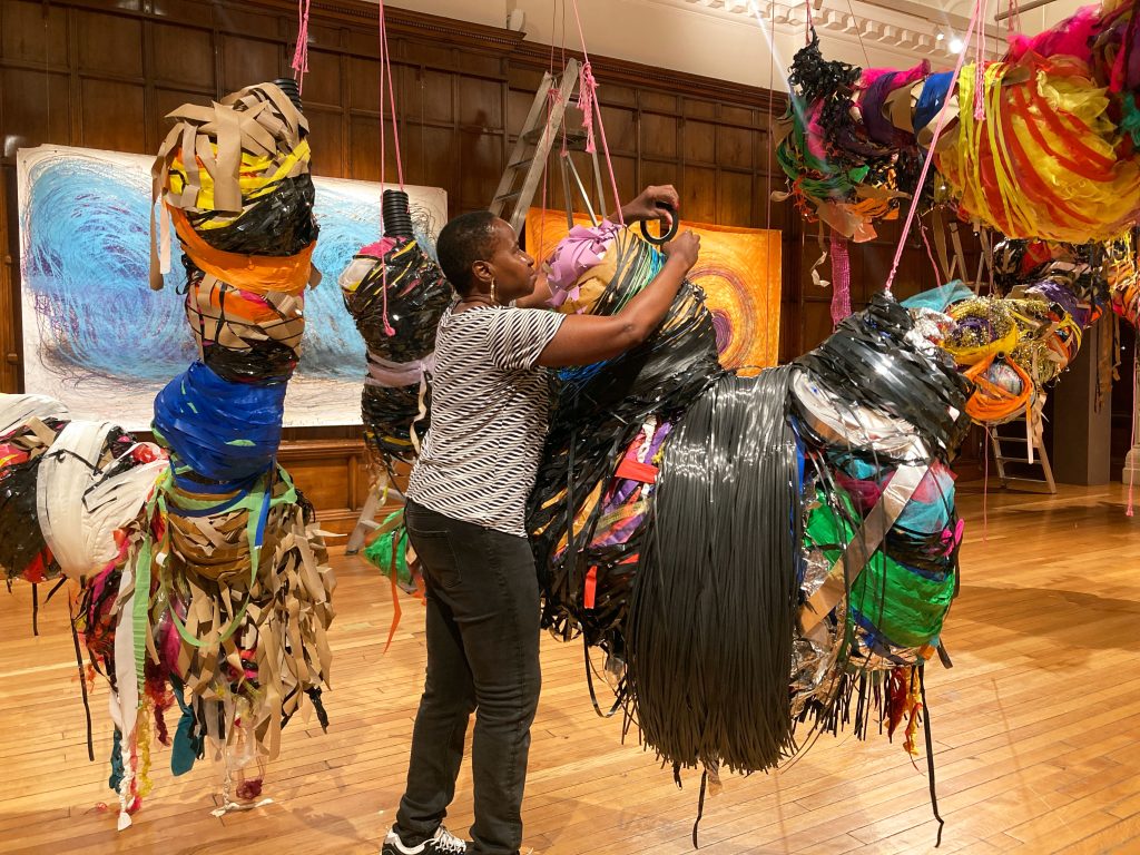 A female artist wraps various materials into floating bundled sculptures in a ornate hall with brown wood walls.