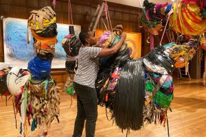 A female artist wraps various materials into floating bundled sculptures in a ornate hall with brown wood walls.