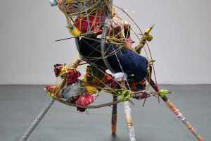 A mixed media, 2ft tall abstract sculpture stands on four legs in an empty large white walled space. The sculpture depicts an animal with colourful recycled materials.