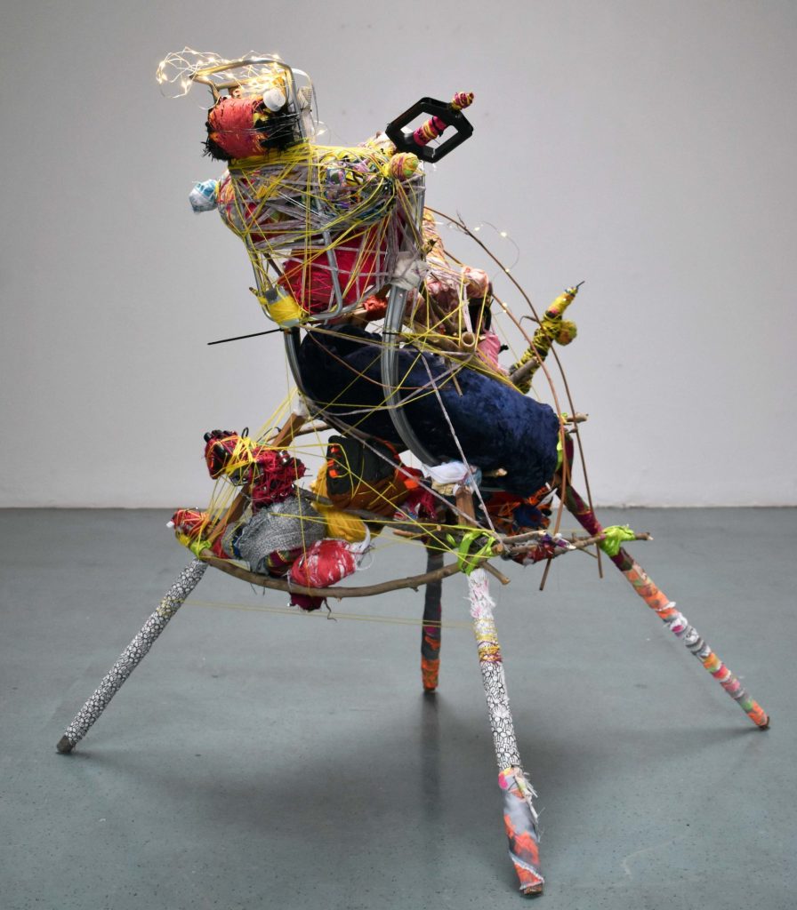 A mixed media, 2ft tall abstract sculpture stands on four legs in an empty large white walled space. The sculpture depicts an animal with colourful recycled materials.