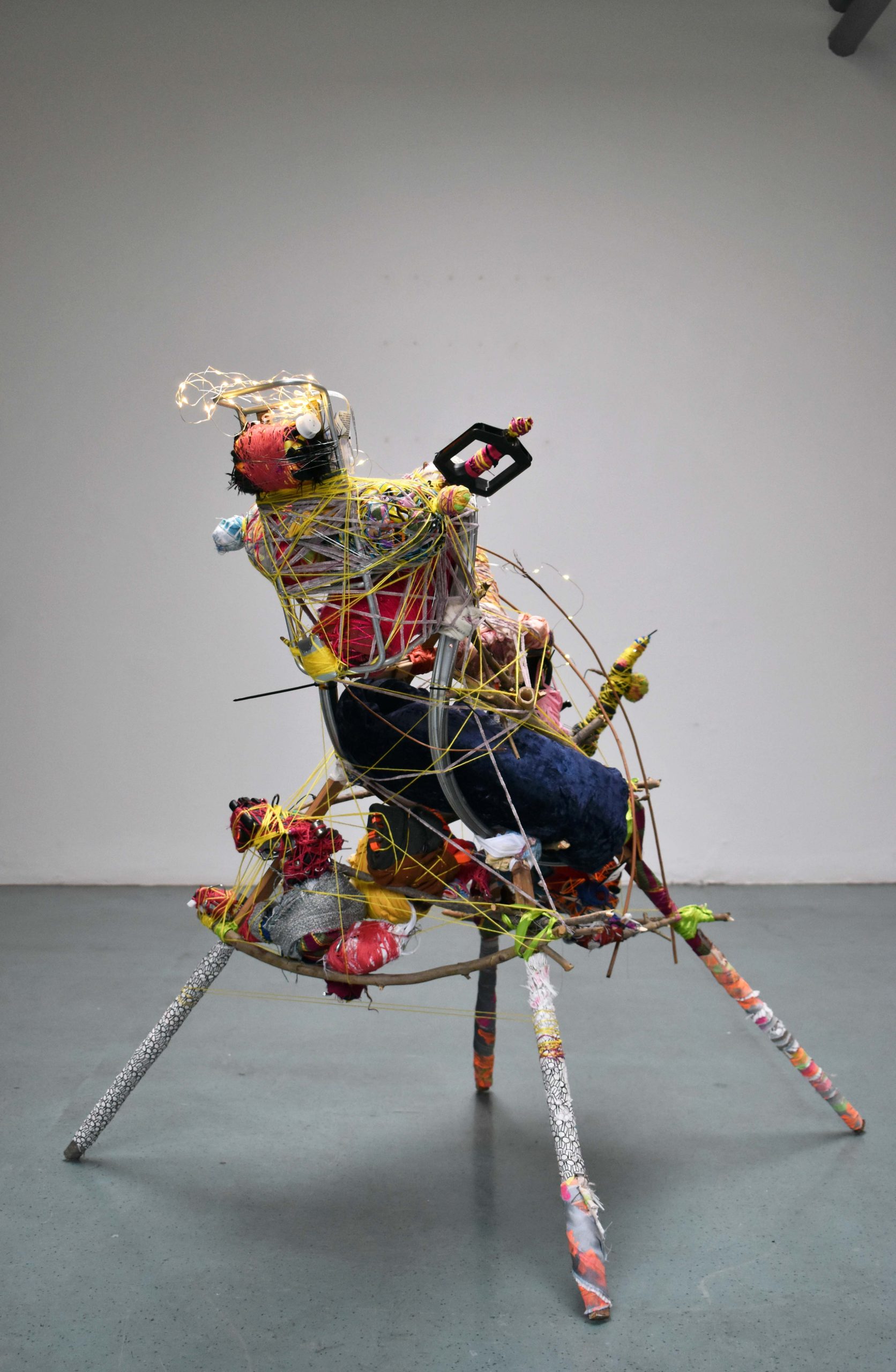 A mixed media, 2ft tall abstract sculpture stands on four legs in an empty large white walled space. The sculpture depicts an animal with colourful recycled materials.