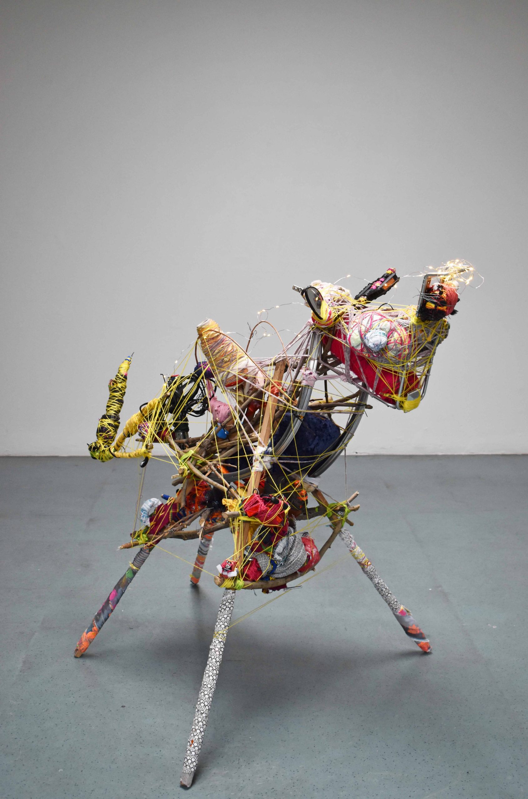 Side angle of a mixed media, 2ft tall abstract sculpture stands on four legs in an empty large white walled space. The sculpture depicts an animal with colourful recycled materials. 