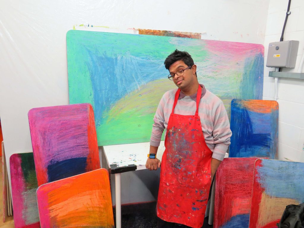 Artist Imran Khan stands in his studio by a desk surrounded by large abstract drawings in pastel.