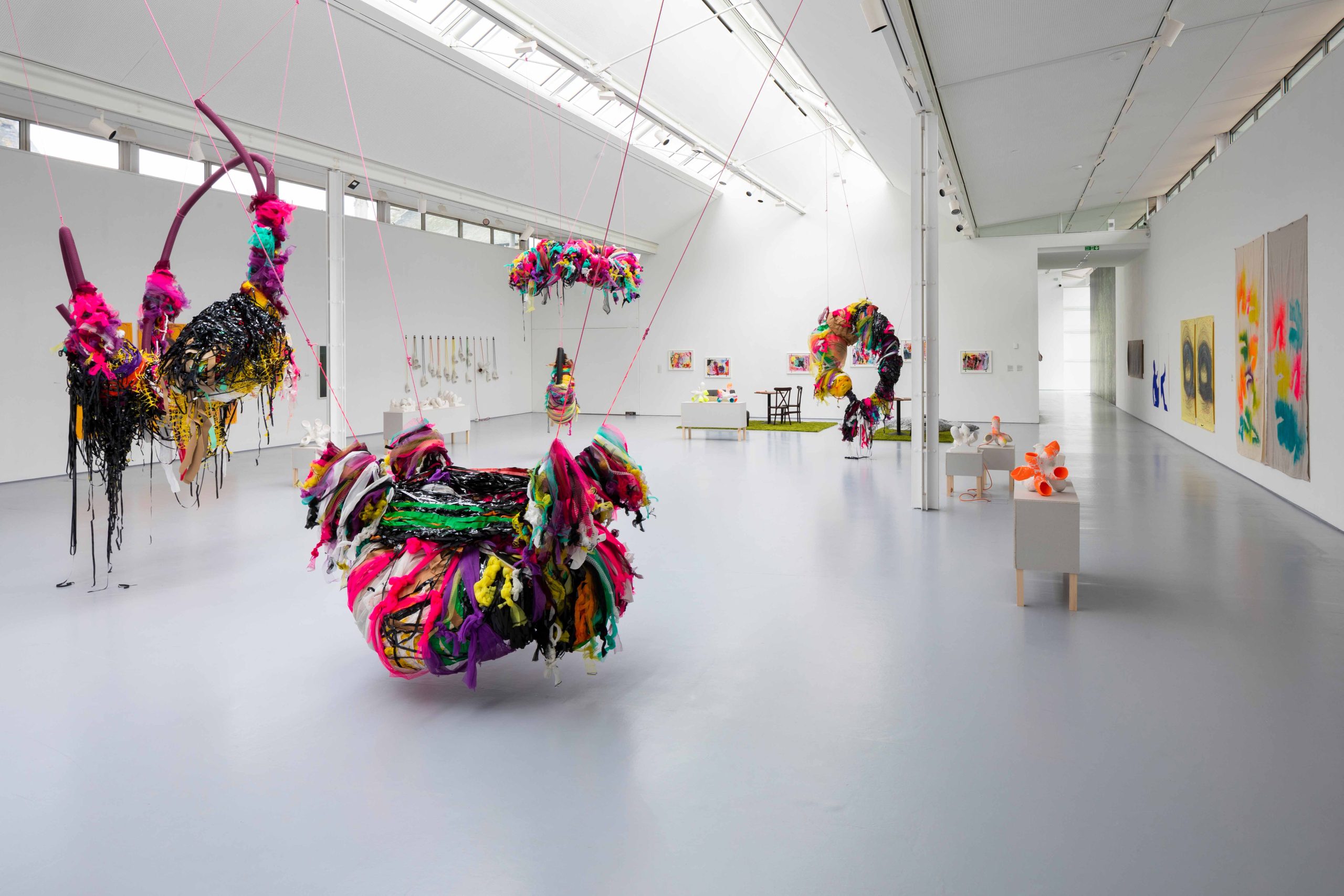 Large, wrapped mixed media sculptures suspend from a ceiling in a large white gallery space with other works displayed on surrounding walls.