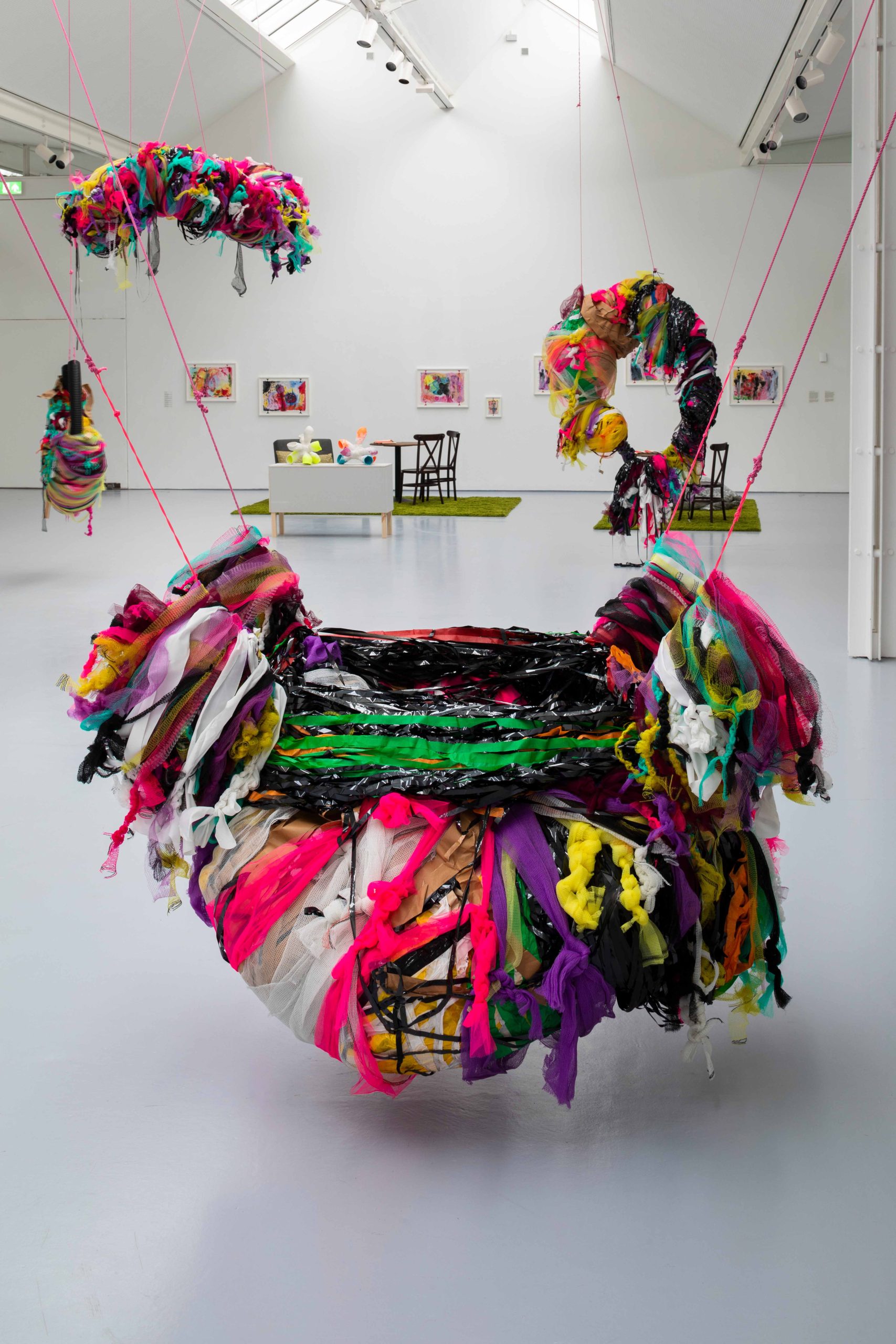 Colourful, large mixed media sculptures hung from a ceiling in a large exhibition space with other works behind.