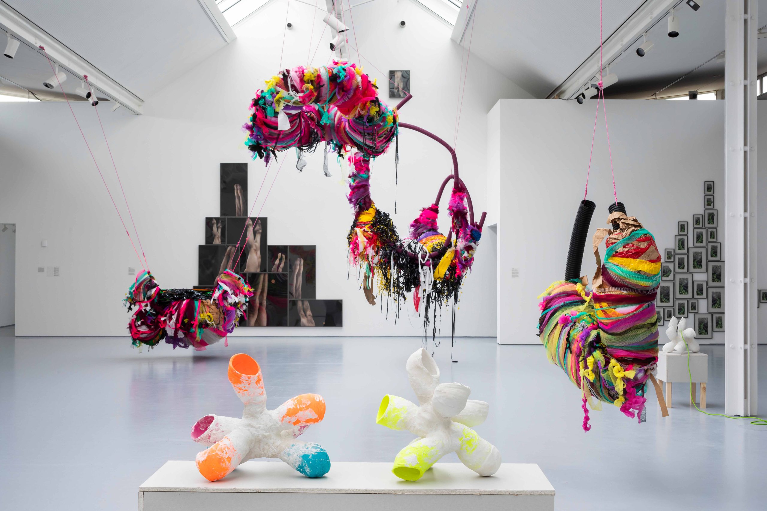 Large, wrapped mixed media sculptures suspend from a ceiling behind small sculptures on a plinth in a large white gallery space with other works displayed on surrounding walls.