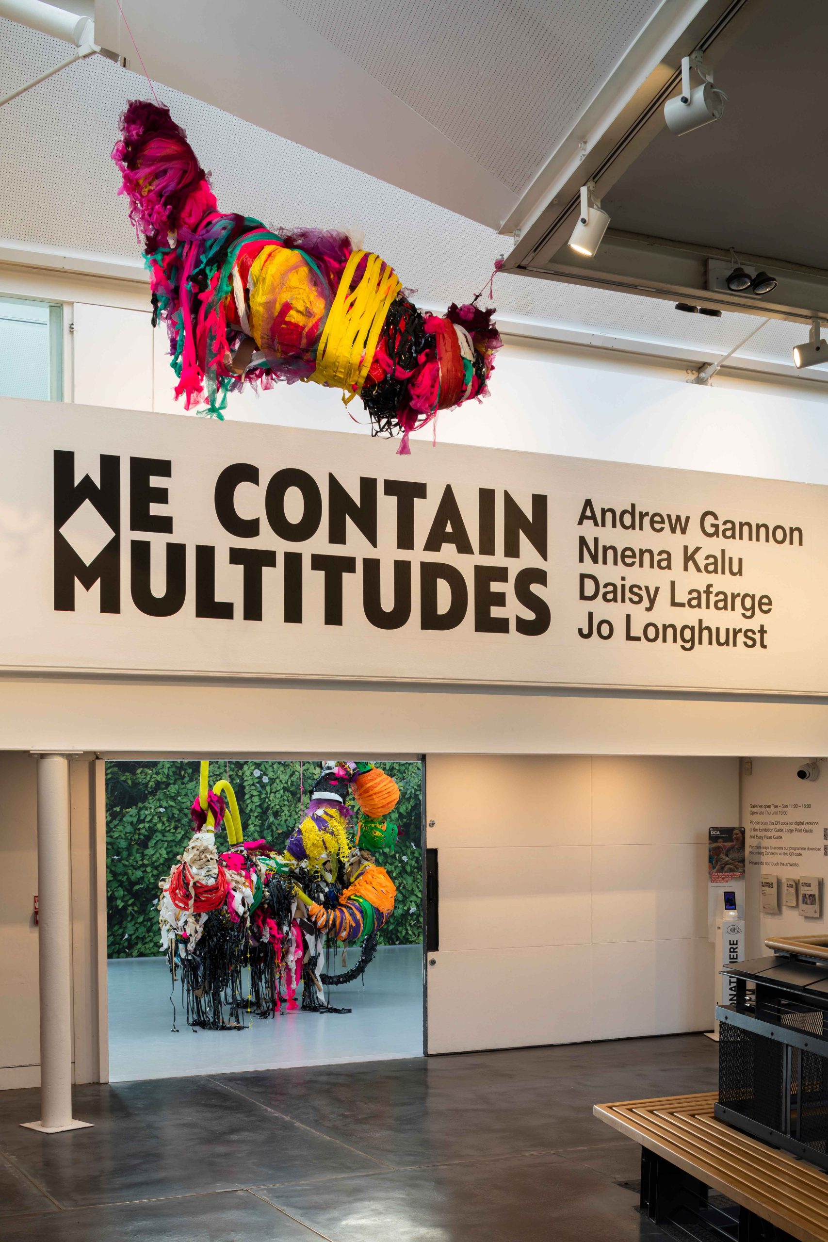 Colourful, large mixed media sculptures hung from a ceiling in a large exhibition space across two floors, large text on wall reads 'WE CONTAIN MULTITUDES'
