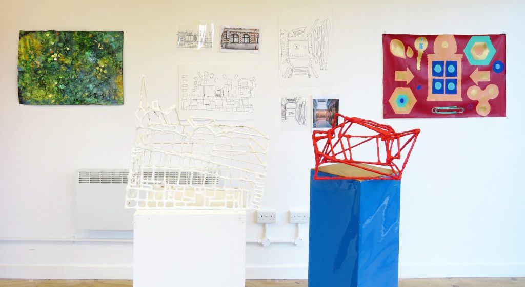 Various mixed media artworks hung on a white wall and on plinths in a gallery space.